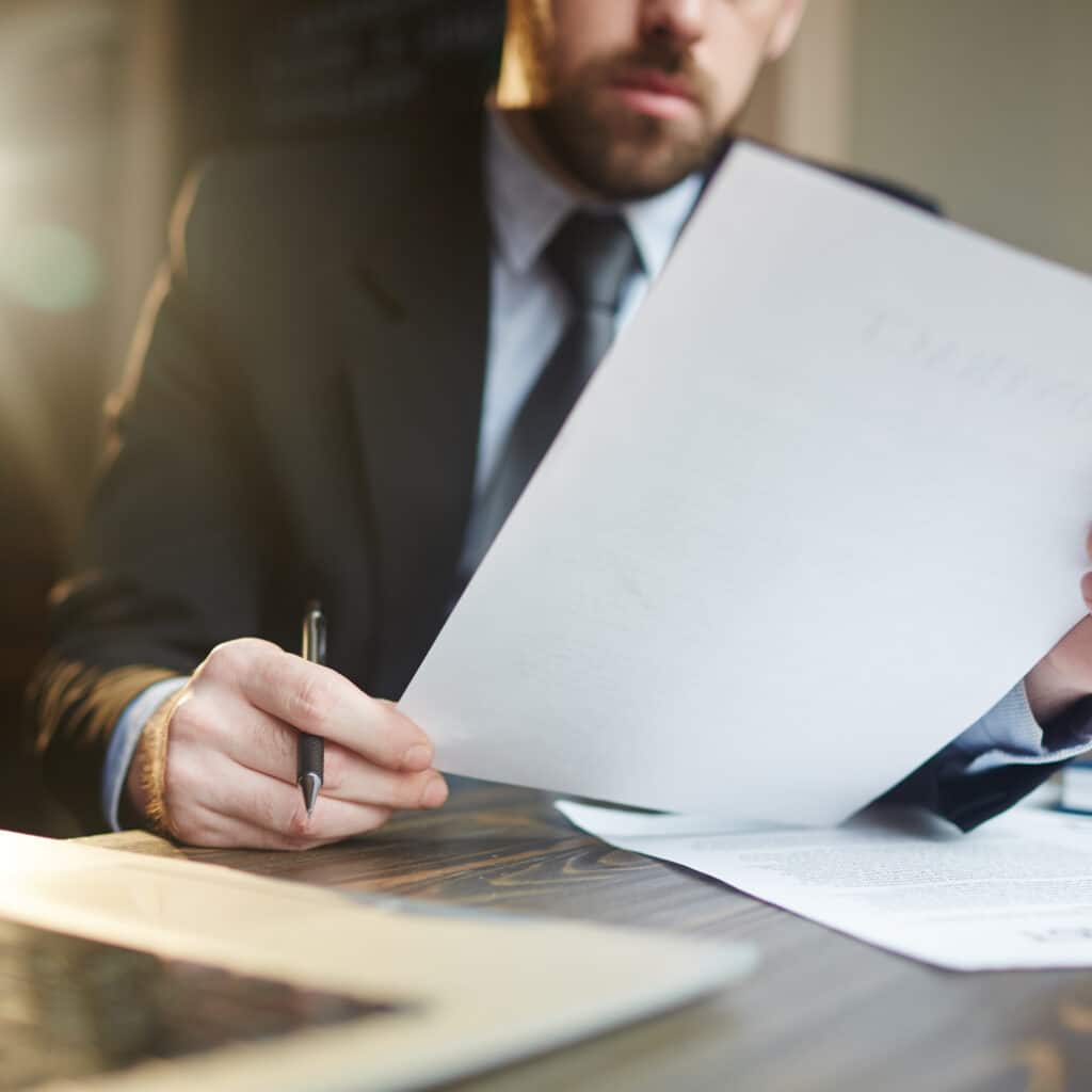 Portrait of modern bearded businessman holding papers in hands, reading and analyzing contract documentation at desk with laptop
